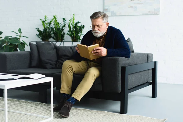 Серйозний бородатий старший чоловік сидить на дивані і читає книгу вдома — стокове фото