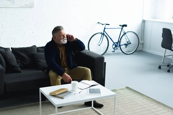 Bearded senior man talking by smartphone while sitting on sofa and looking away at home — Stock Photo