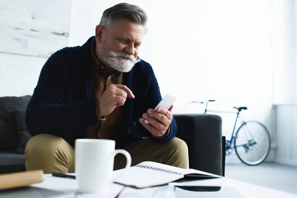 Hombre mayor sonriente guapo usando teléfono inteligente mientras está sentado en el sofá en casa - foto de stock