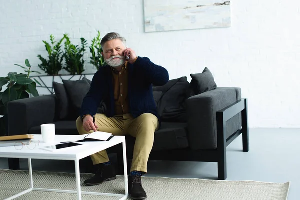 Sonriente barbudo hombre mayor sentado en el sofá y hablando por teléfono inteligente - foto de stock