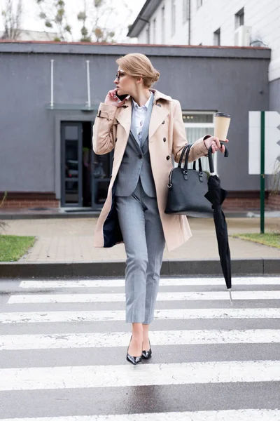 Mujer de negocios con café para ir y paraguas hablando en el teléfono inteligente mientras cruza la calle en la calle - foto de stock