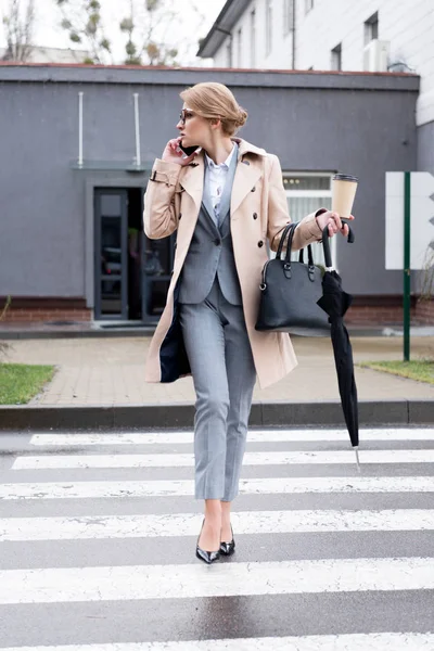 Businesswoman with coffee to go and umbrella talking on smartphone while crossing road on street — Stock Photo