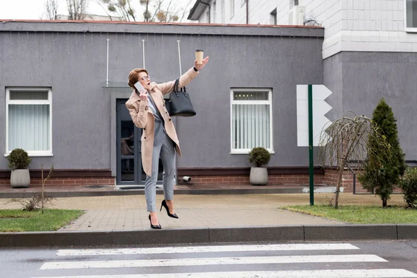 Mujer de negocios con café para ir a hablar en el teléfono inteligente y llamar a taxi en la calle - foto de stock