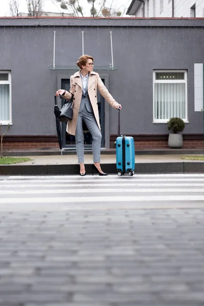 Бизнесвумен с чемоданом и зонтиком ждет такси на улице — стоковое фото