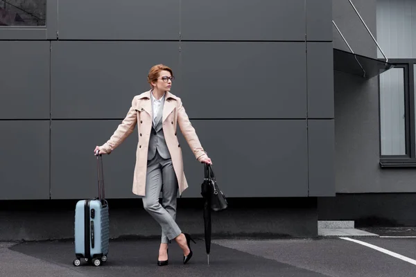 Elegante mujer de negocios en abrigo con maleta esperando taxi en la calle - foto de stock