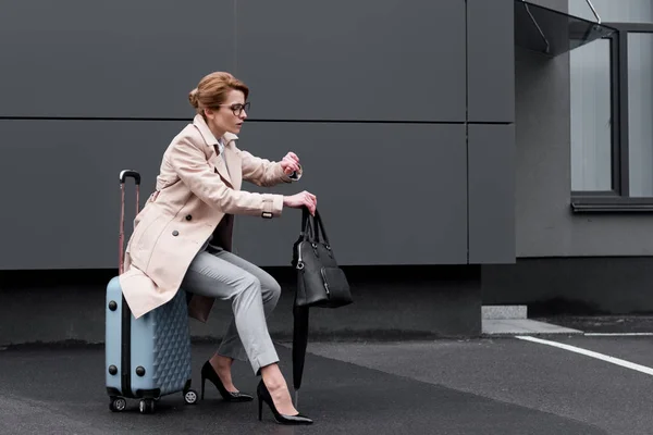 Mujer de negocios con un abrigo elegante sentado en la maleta mientras espera un taxi en la calle — Stock Photo