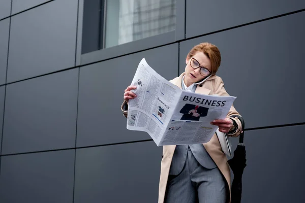 Business woman talking on smartphone while reading newspaper on street — стоковое фото