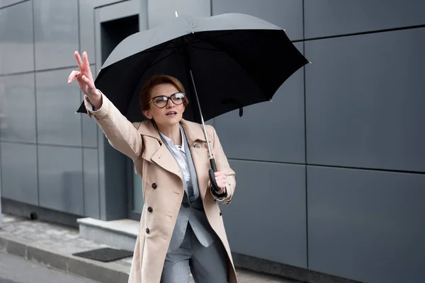 Geschäftsfrau im schicken Mantel mit Regenschirm ruft Taxi auf der Straße — Stockfoto