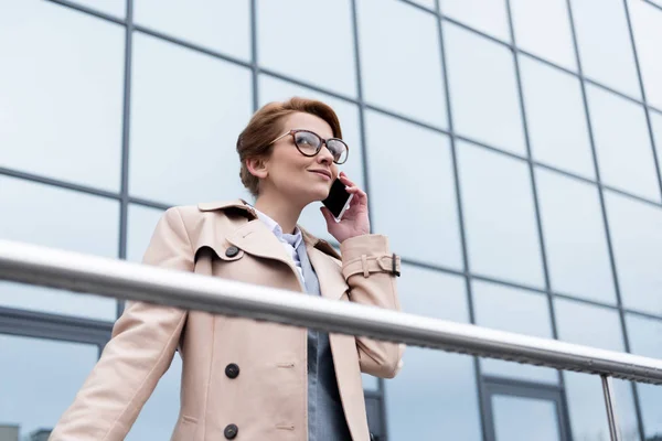 Vista de ángulo bajo de la mujer de negocios sonriente hablando en el teléfono inteligente en la calle - foto de stock