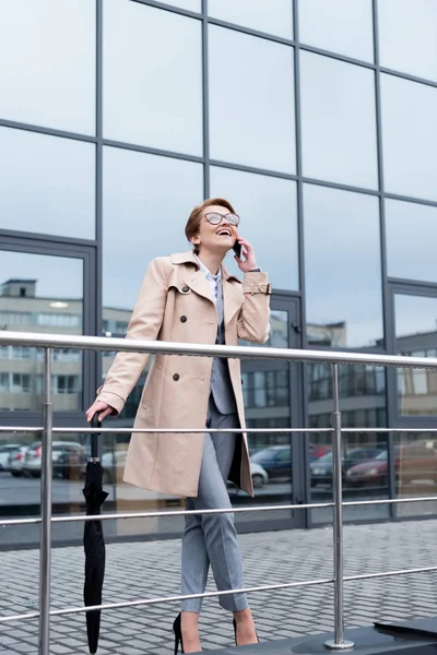 Happy businesswoman with umbrella talking on smartphone on street — Stock Photo
