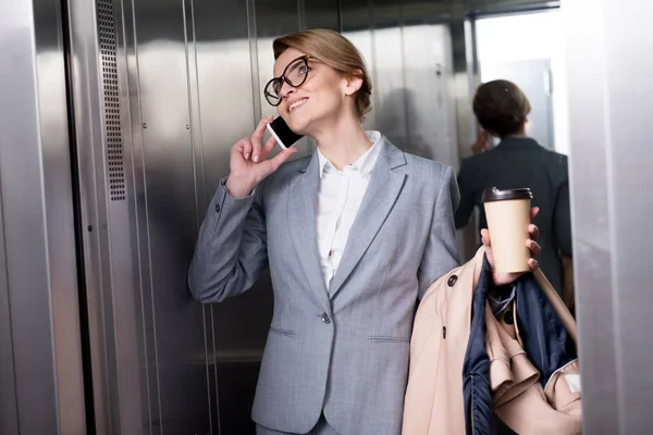Lächelnde Geschäftsfrau im Anzug spricht im Fahrstuhl auf Smartphone — Stockfoto