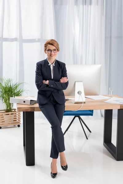 Smiling businesswoman with arms crossed leaning on table in office — Stock Photo