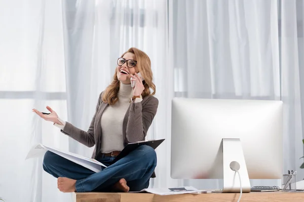 Счастливая деловая женщина с бумагами разговаривая на смартфоне, сидя на столе в офисе — стоковое фото