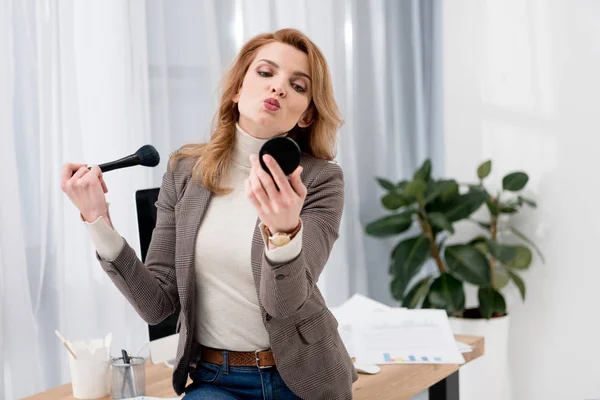 Стильная деловая женщина, смотрящая в зеркало при нанесении макияжа на рабочем месте в офисе — стоковое фото