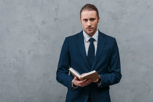 Young handsome businessman with book looking at camera in front of concrete wall — Stock Photo