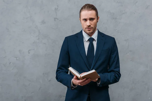 Joven hombre de negocios guapo con libro mirando a la cámara delante de la pared de hormigón — Stock Photo