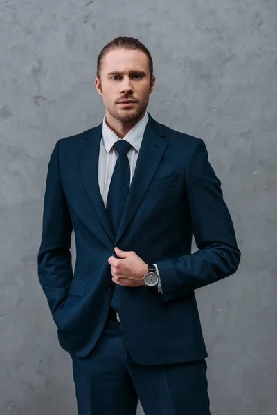 Joven hombre de negocios guapo en traje elegante delante de la pared de hormigón mirando a la cámara — Stock Photo