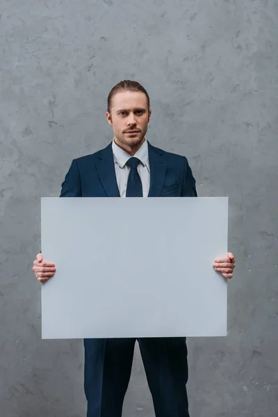 Young confident businessman holding blank board in front of concrete wall — Stock Photo
