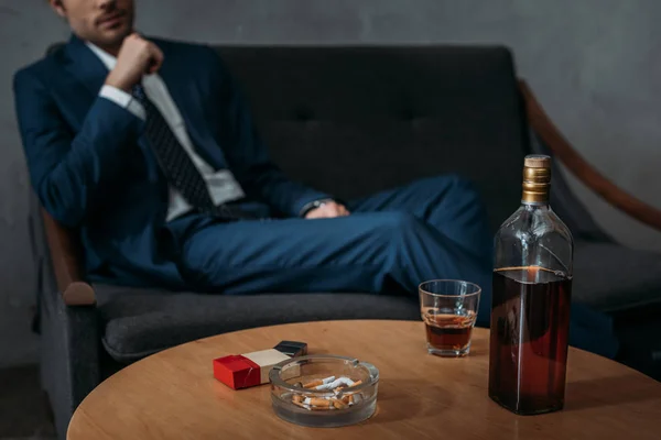 Recortado tiro de hombre de negocios sentado en el sofá delante de la mesa con botella de whisky - foto de stock