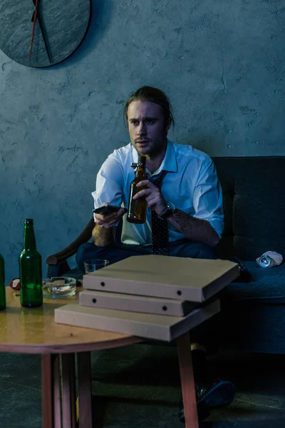Молодой алкоголик в белой рубашке смотрит телевизор и пьет пиво после работы на диване — стоковое фото