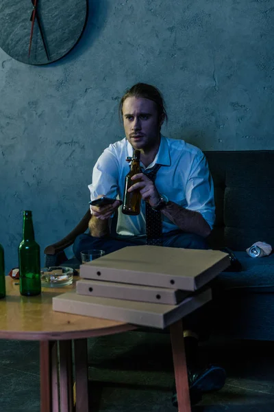 Young alcohol addicted man in white shirt watching tv and drinking beer after work on couch — Stock Photo