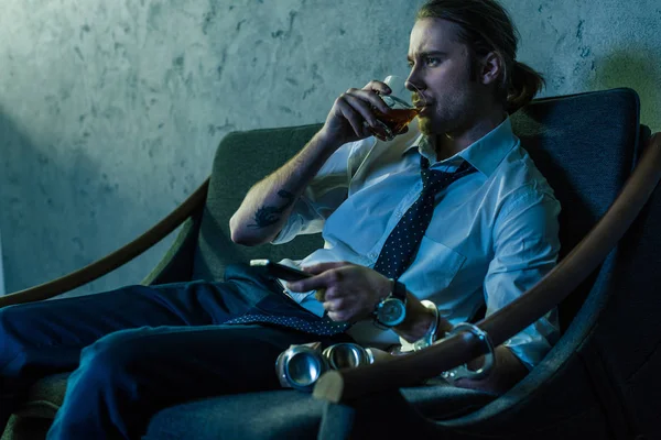 Hombre adicto al alcohol en camisa blanca viendo la televisión y bebiendo whisky después del trabajo - foto de stock