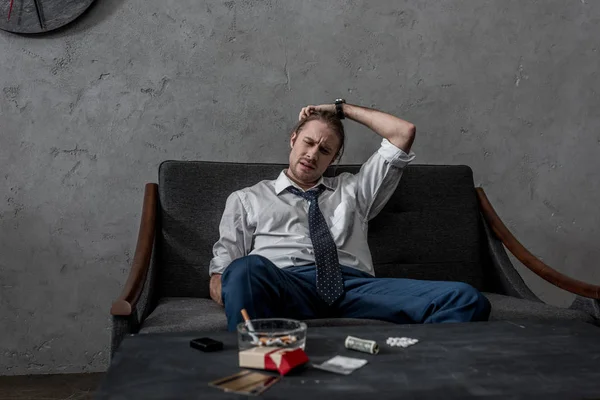 Hombre de negocios deprimido con adicción a las drogas sentado en el sofá frente a la mesa con drogas - foto de stock