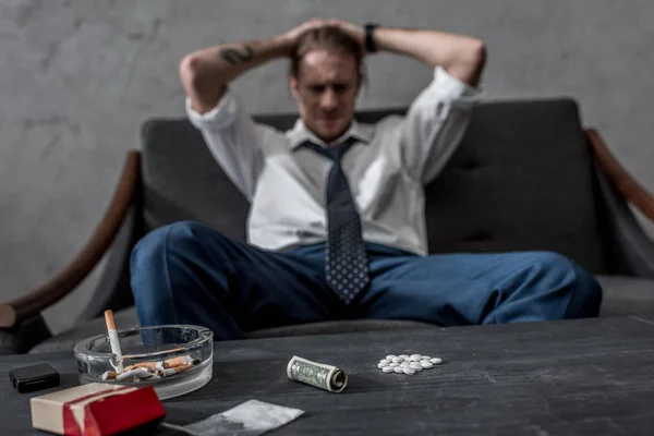 Hombre de negocios deprimido con adicción a las drogas sentado en el sofá frente a la mesa con píldoras mdma - foto de stock