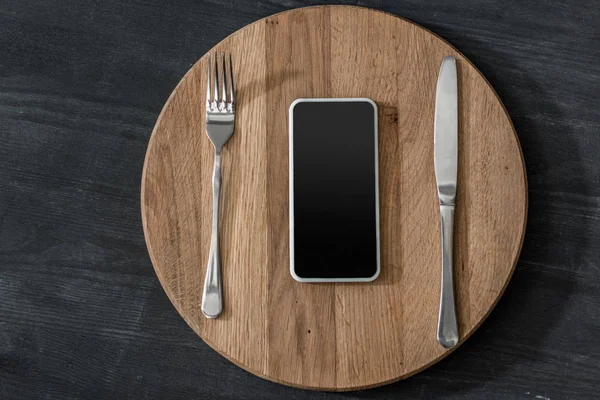 Top view of smartphone lying on wooden board with cutlery, phone addiction concept — Stock Photo