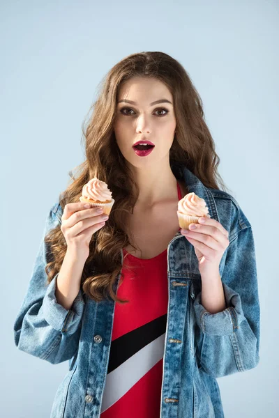Chica atractiva sorprendida sosteniendo cupcakes y mirando a la cámara aislada en gris - foto de stock