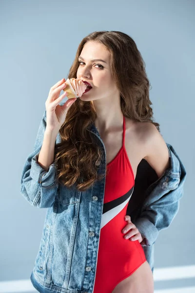 Sexy chica mordiendo cupcake y mirando a la cámara en gris - foto de stock