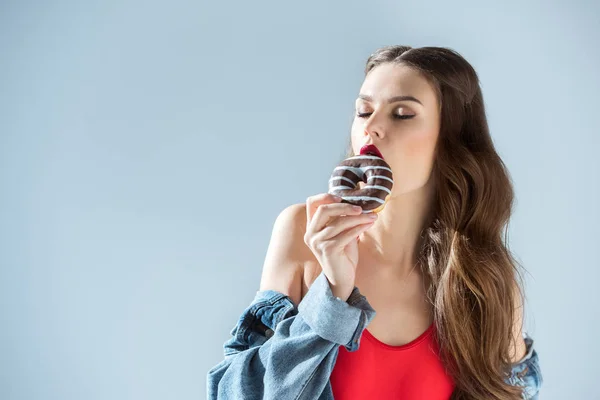 Sexy chica en rojo traje de baño mordiendo donut con los ojos cerrados aislado en gris - foto de stock