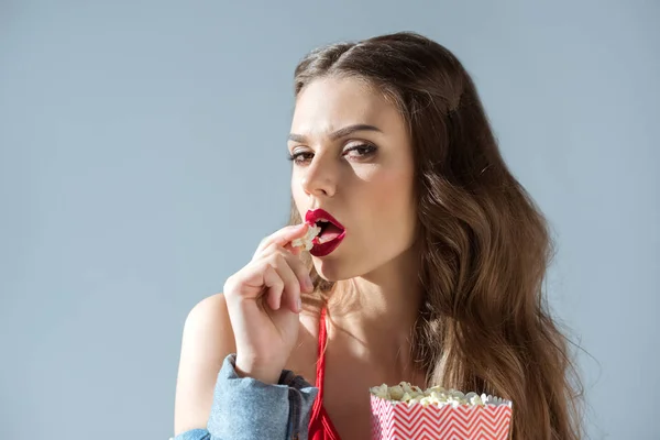 Соблазнительная девушка ест попкорн и смотрит на камеру, изолированную от серого — стоковое фото