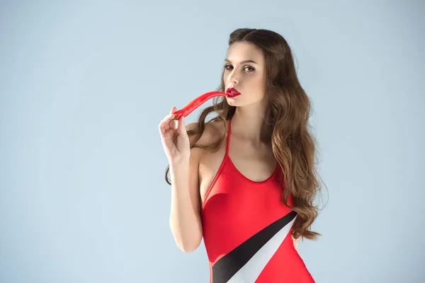 Seductive woman in red swimsuit eating red chili pepper and looking at camera isolated on grey — Stock Photo