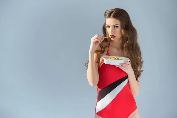 Sexy girl in red swimsuit eating french fries and looking at camera isolated on grey — Stock Photo