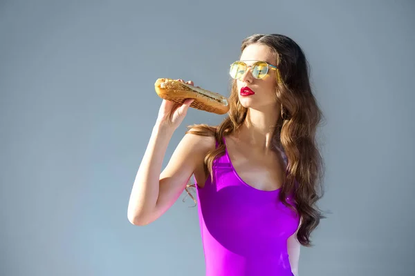 Sexy attractive girl in ultra violet swimsuit looking at hot dog isolated on grey — Stock Photo