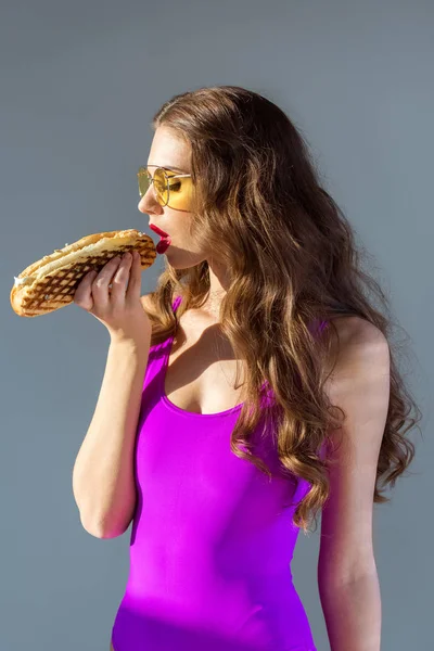 Sexy attractive girl in ultra violet swimsuit eating hot dog isolated on grey — Stock Photo