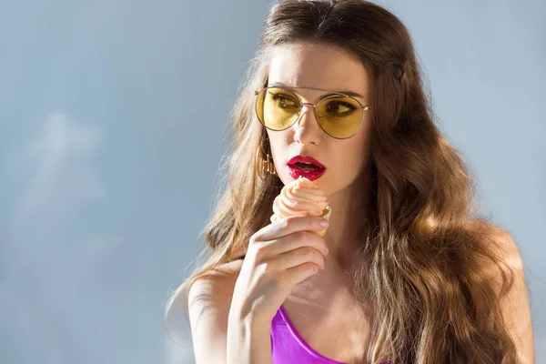 Sexy attrayant fille en maillot de bain ultra violet et lunettes de soleil manger cupcake sur gris — Photo de stock