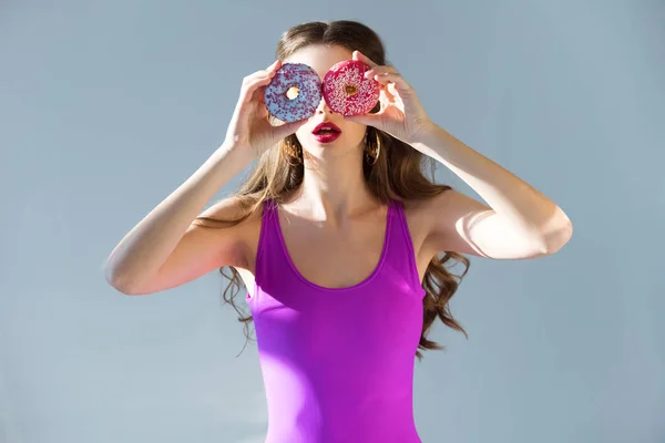 Sexy chica atractiva en traje de baño ultra violeta cubriendo los ojos con rosquillas aisladas en gris - foto de stock