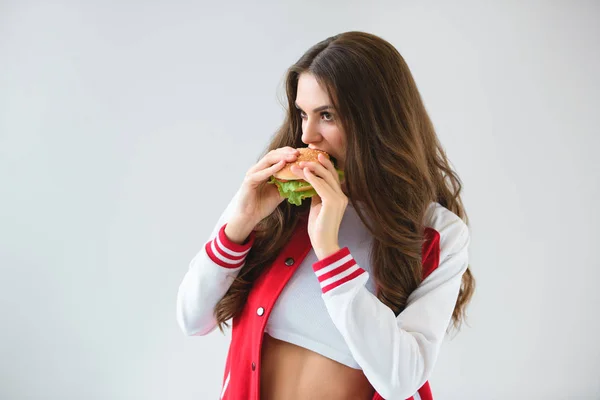 Menina sedutora em jaqueta de beisebol comer hambúrguer isolado no branco — Fotografia de Stock