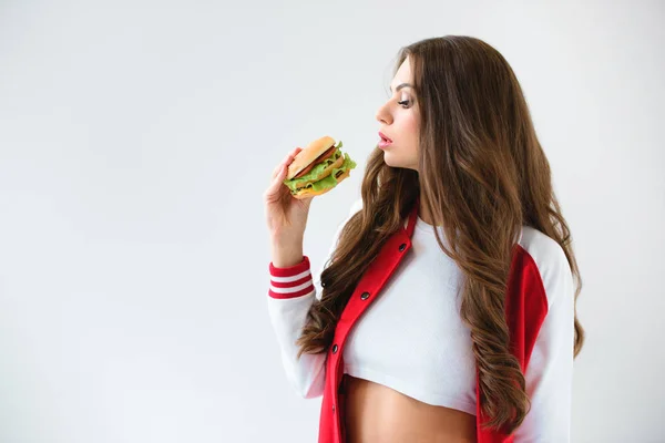 Vista lateral da menina sedutora em jaqueta de beisebol olhando para hambúrguer isolado no branco — Fotografia de Stock