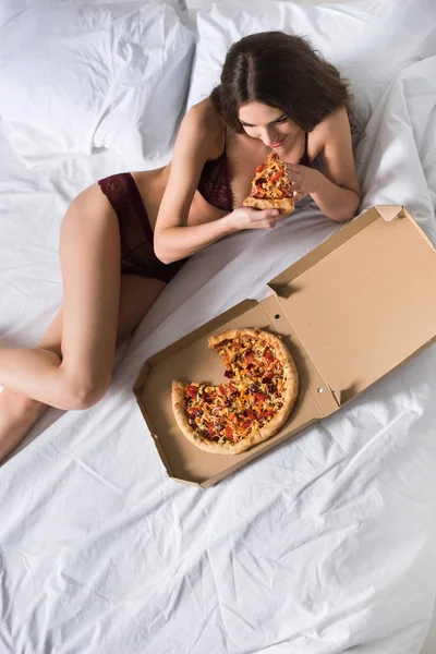 Vista de ángulo alto de la mujer seductora acostada en ropa interior de encaje en la cama y comer pizza - foto de stock