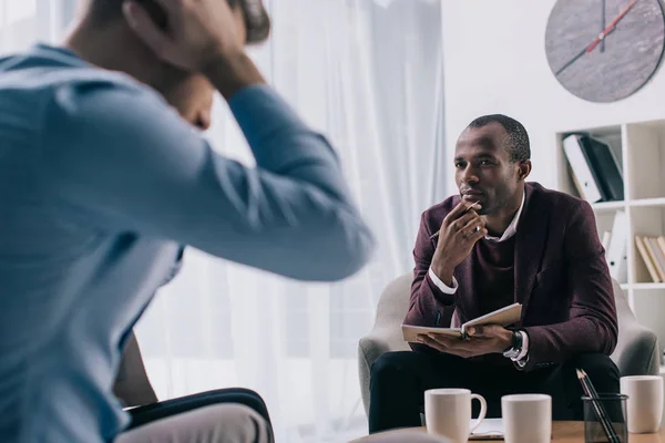 Frustrierter junger Mann sitzt auf Sofa und afrikanisch-amerikanischer Psychiater im Büro — Stockfoto