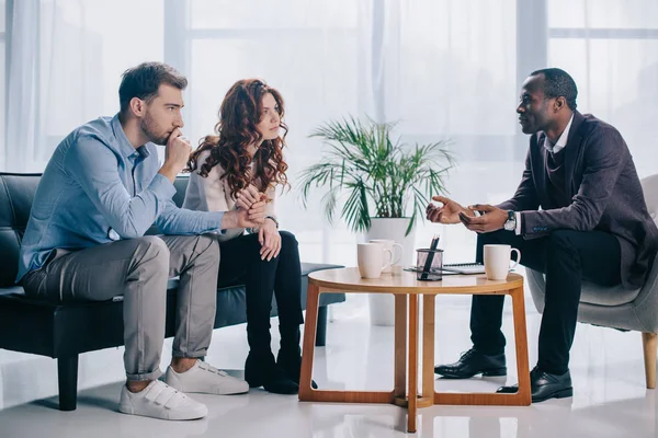 Un psychiatre afro-américain souriant parle à un jeune couple au bureau — Photo de stock