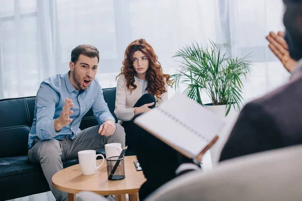 Wütender junger Mann mit Freundin sitzt auf Sofa und streitet mit Berater — Stockfoto