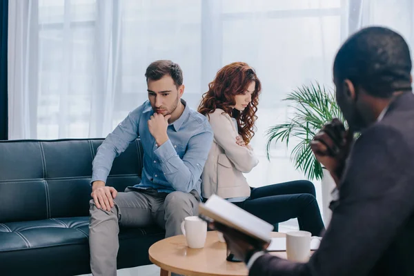 Divorcing couple sitting back to back on sofa and african american counselor with textbook — Stock Photo