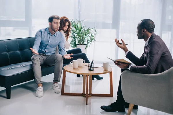 Joven enojado con novia sentado en el sofá y discutiendo con el consejero — Stock Photo