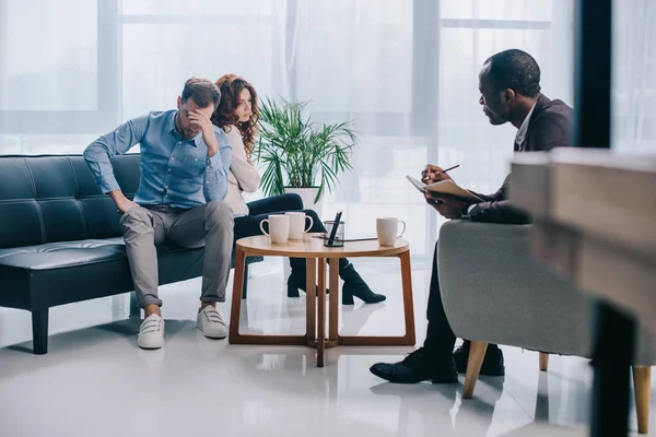 Africano conselheiro americano escrevendo no livro enquanto chateado jovem casal sentado no sofá — Stock Photo