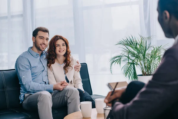 Lächelndes junges Paar auf Sofa sitzend und mit Bleistift in der Hand mit Berater im Gespräch — Stockfoto