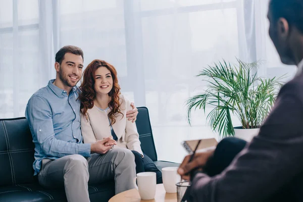 Sorrindo jovem casal sentado no sofá e conversando com o conselheiro com lápis na mão — Fotografia de Stock